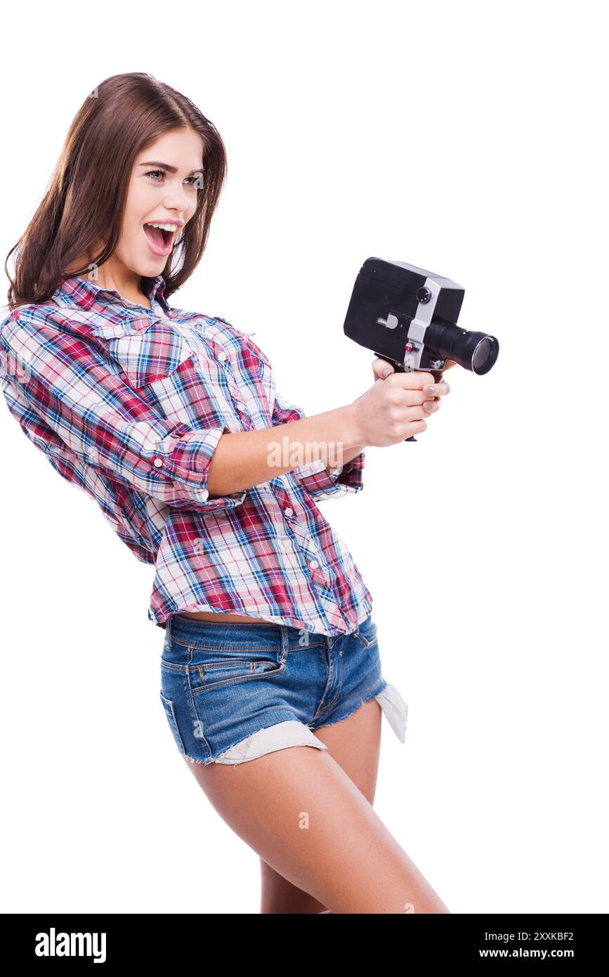 Telecamera cinematografica come arma. Giovane donna allegra che tiene in mano la macchina fotografica mentre è in piedi su sfondo bianco Foto Stock