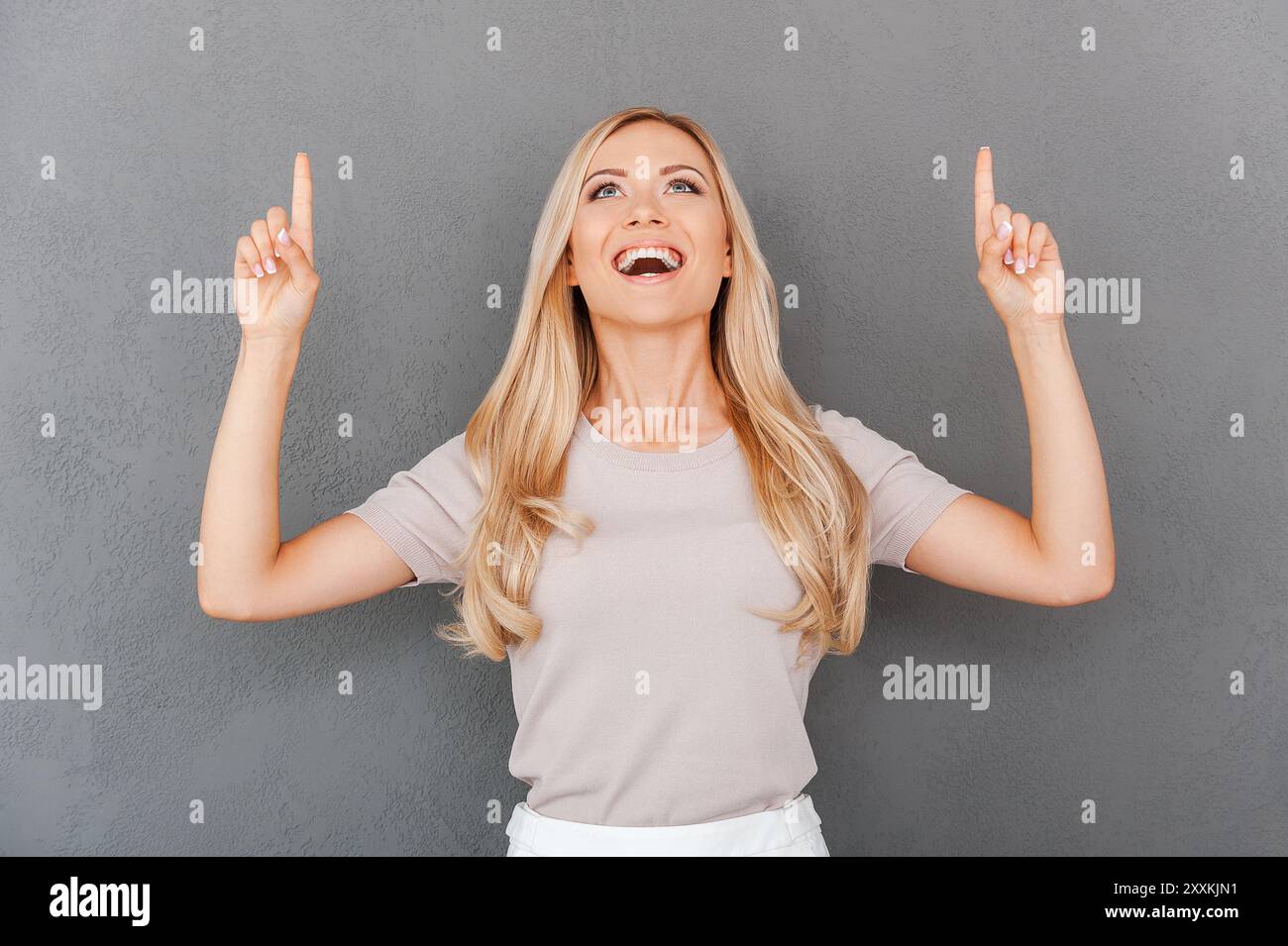 Portare il business a nuovi livelli. Giovane e allegra donna bionda che punta in alto e sorride mentre sta in piedi su sfondo grigio Foto Stock