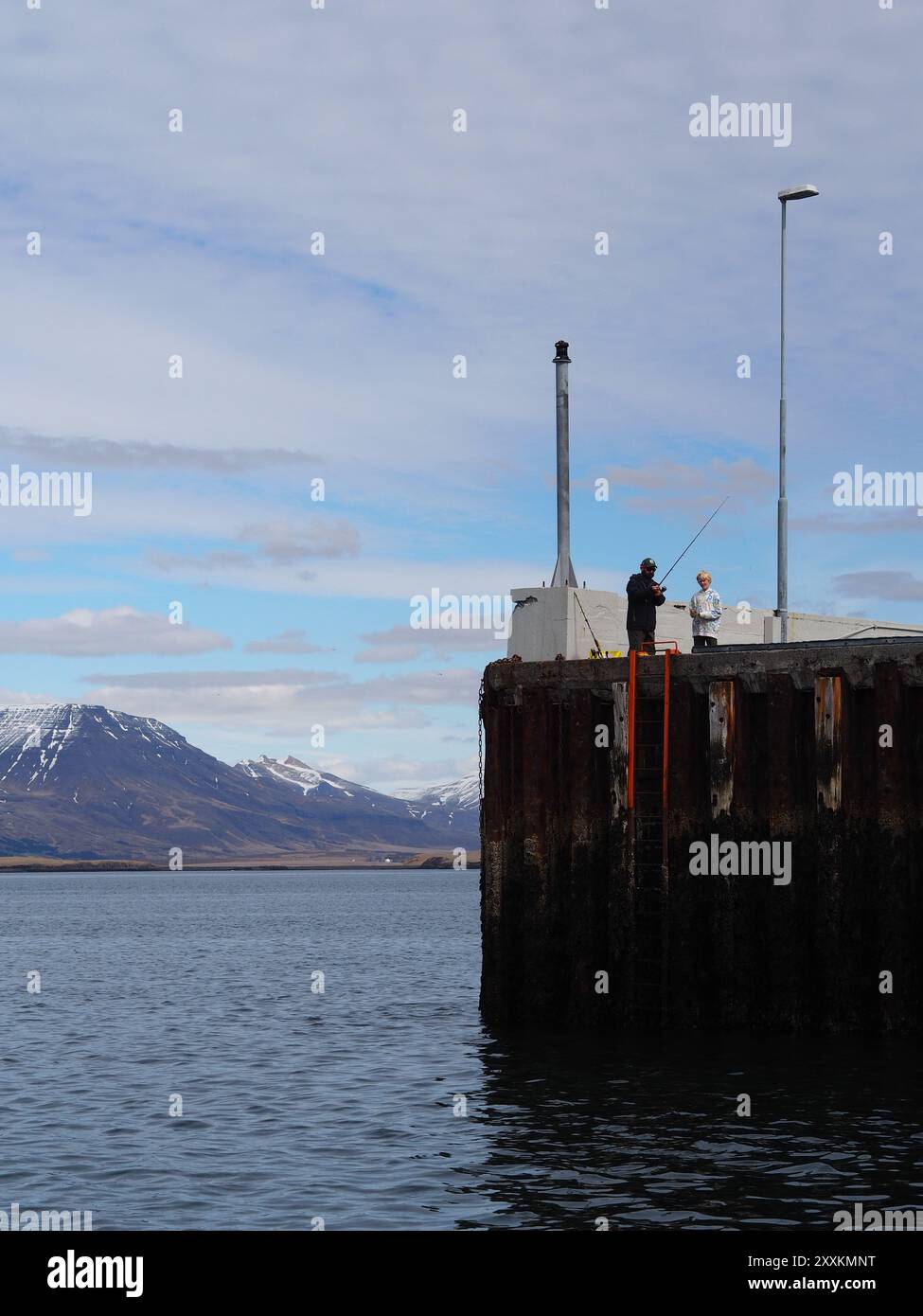 Due persone stanno su un molo, pescando e godendosi l'ambiente tranquillo, con uno splendido sfondo di montagna e un cielo blu parzialmente nuvoloso che esalta il Foto Stock