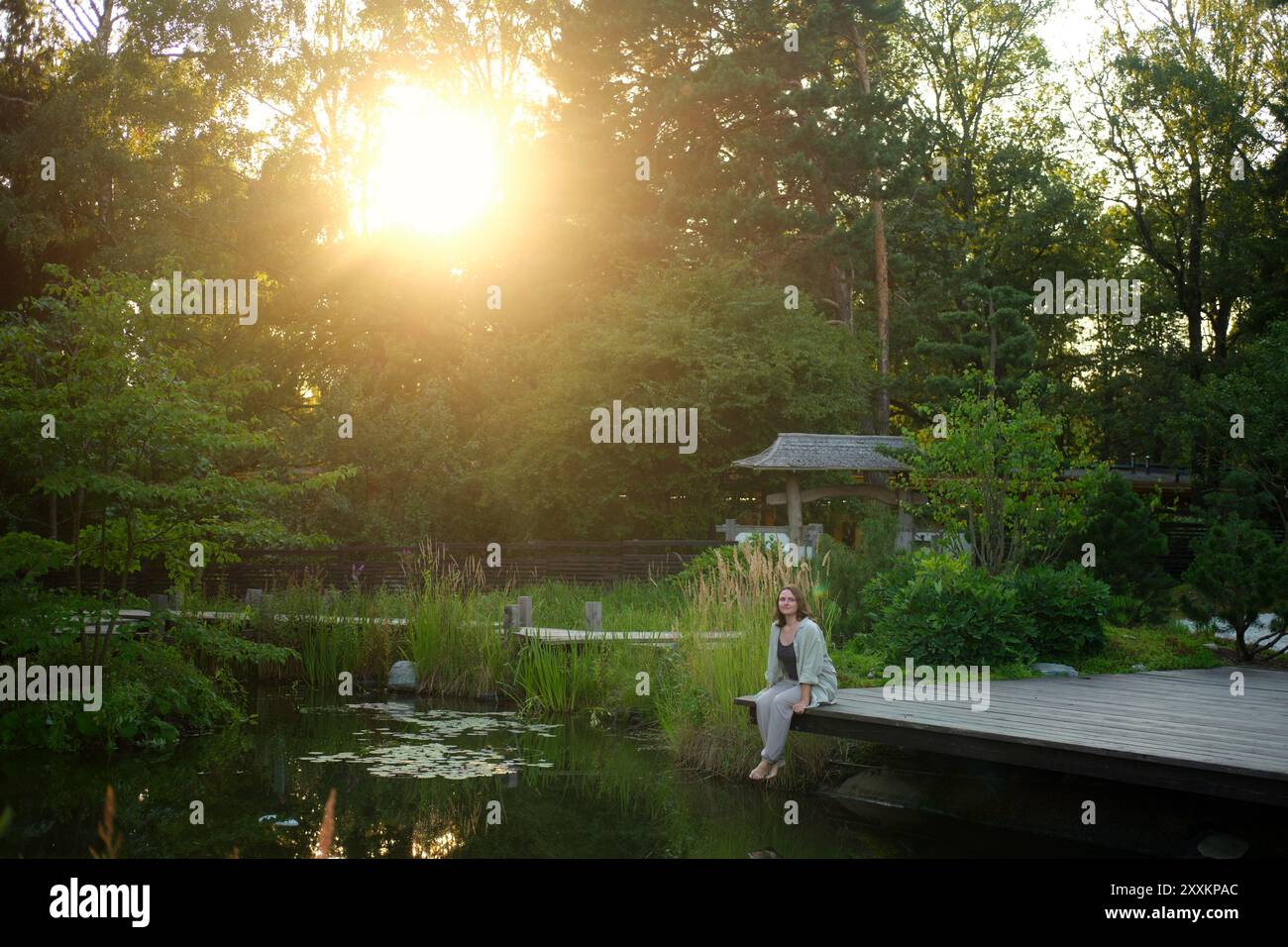 Donna seduta su un ponte di legno vicino a uno stagno con vegetazione lussureggiante e tradizionale cancello giapponese al tramonto. Concetto di natura e relax. Foto Stock