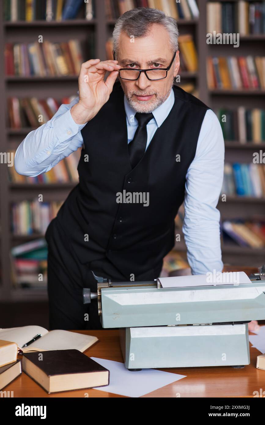 Intelligente e sicuro di sé. Uomo anziano con capelli grigi sicuri in abbigliamento formale appoggiato al tavolo con una macchina da scrivere e una libreria sullo sfondo Foto Stock