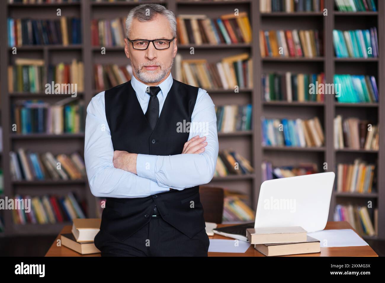 Professore fiducioso. Capelli grigi sicuri uomo anziano in abbigliamento formale che tiene le braccia incrociate e guarda la fotocamera mentre si appoggia al tavolo e con i libri Foto Stock