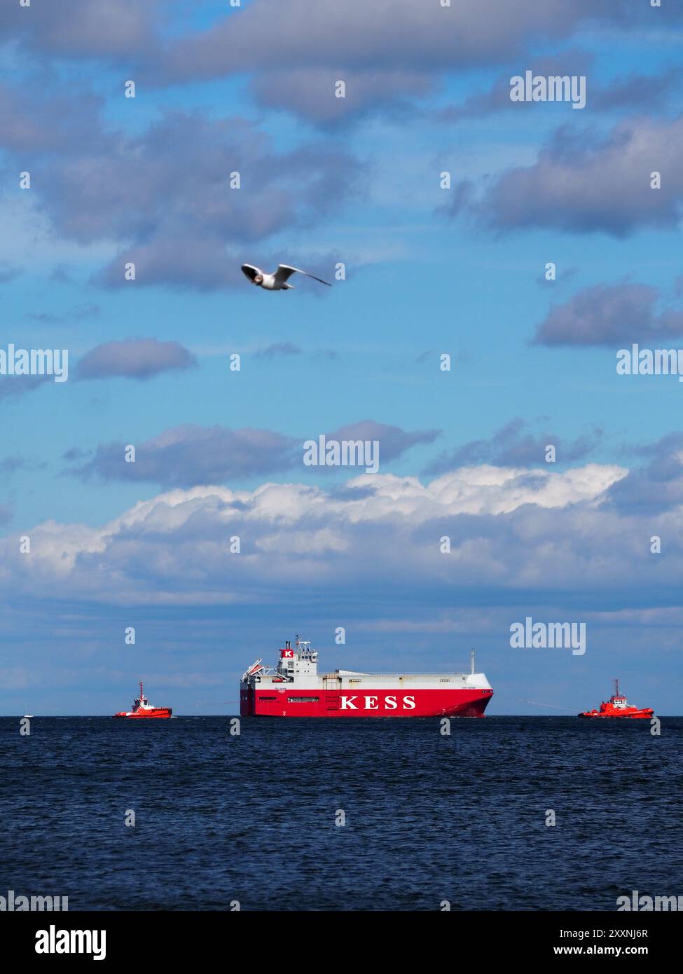 Danzica, Polonia - 13 luglio 2024: Una massiccia nave da carico rossa KESS naviga attraverso l'oceano, accompagnata da due barche più piccole, sotto un cielo azzurro limpido con sc Foto Stock