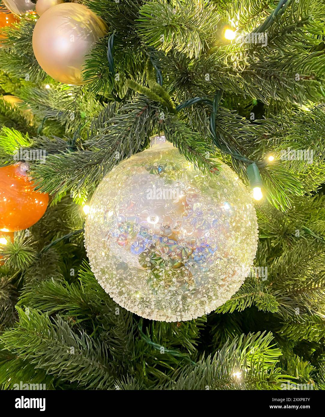 Splendido ornamento a forma di palla di albero di Natale in vetro in primo piano, appeso a foglie di pino verde con minuscole luci, ornamenti oro e arancio Foto Stock