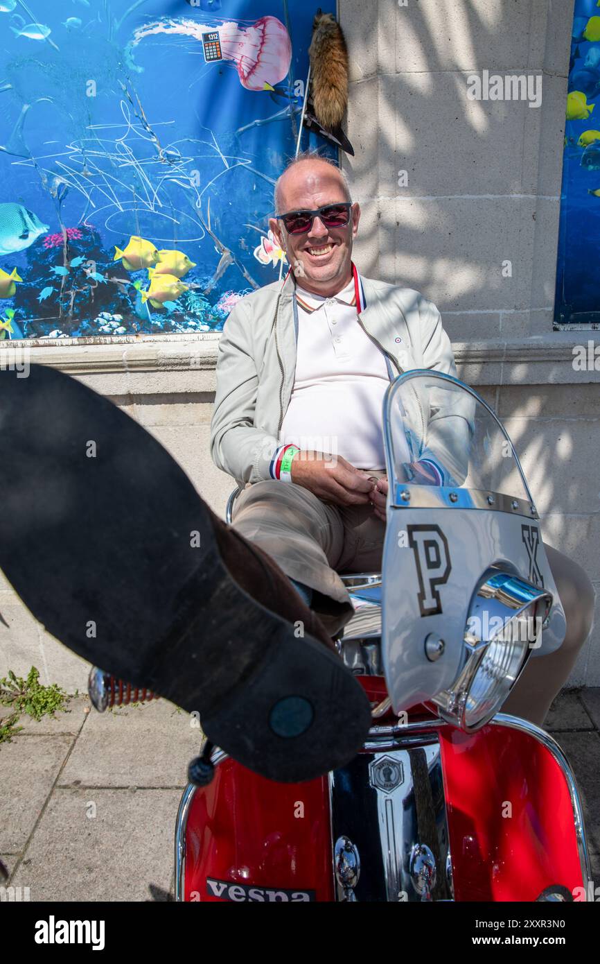 Un Mod siede su uno scooter con i piedi in alto. Migliaia di persone partecipano all'annuale Mod Weekender che si tiene nella cittadina costiera inglese di Brighton. Quest'anno celebra il 60° anniversario della battaglia di Brighton, una delle più famose battaglie di Brighton tra i Mods e i Rockers. Negli anni '1950 e '1960 le due sottoculture giovanili si sono riunite nelle località balneari dell'Inghilterra meridionale per combattere le battaglie tra loro e la polizia. Ciò causò una protesta morale pubblica da parte di una società conservatrice del dopoguerra che vide gli autori arrestati, processati e imprigionati. Mentre la cultura Rocker è meno Foto Stock