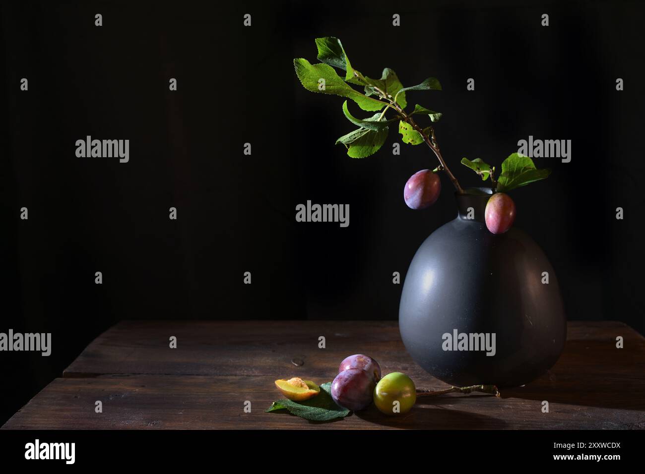 Ramoscello di prugna con frutta e foglie in un vaso e alcune prugne su un rustico tavolo di legno su sfondo nero, natura morta cibo scuro e moody, copia S. Foto Stock