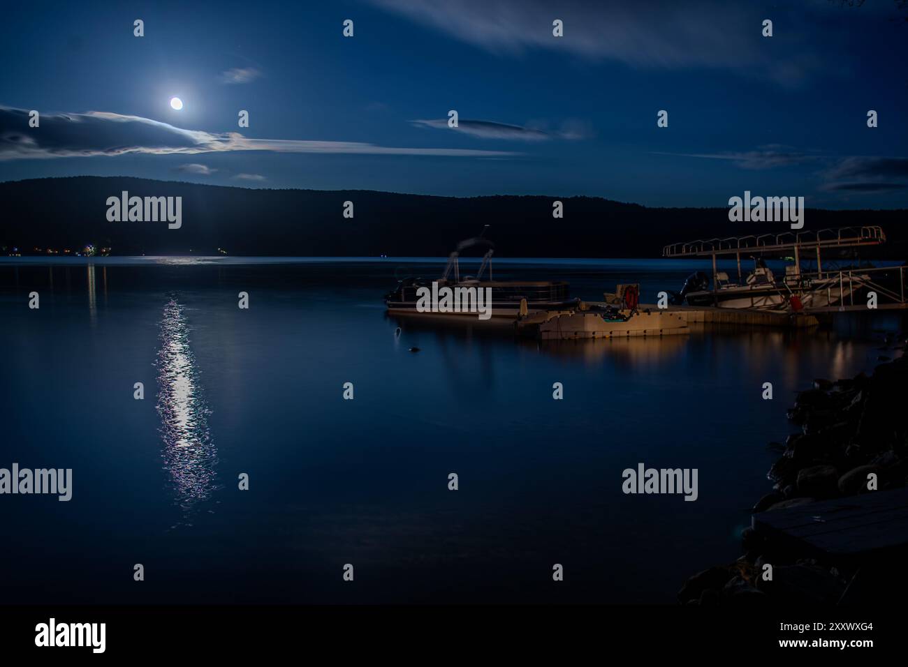 Una luna piena sorge sul lago Otsego a Cooperstown, New York, e il suo riflesso risplende sull'acqua mentre un molo con le barche riposa pacificamente lungo il fiume Foto Stock