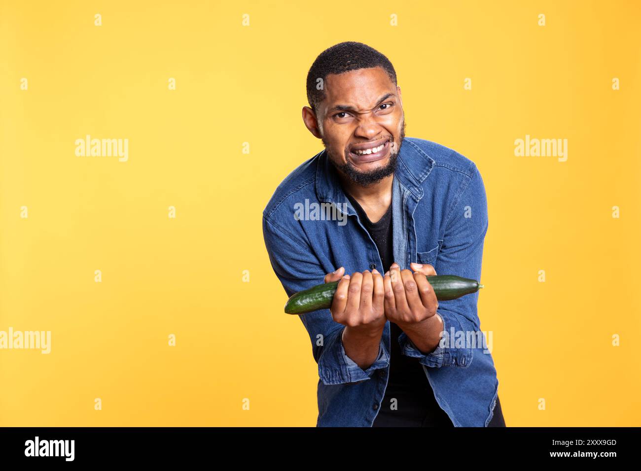 Ragazzo afroamericano che si prende gioco con un cetriolo, comportandosi come se facesse sollevamento pesi su sfondo giallo. Adulto motivato e felice che si diverte con verdure fresche. Foto Stock