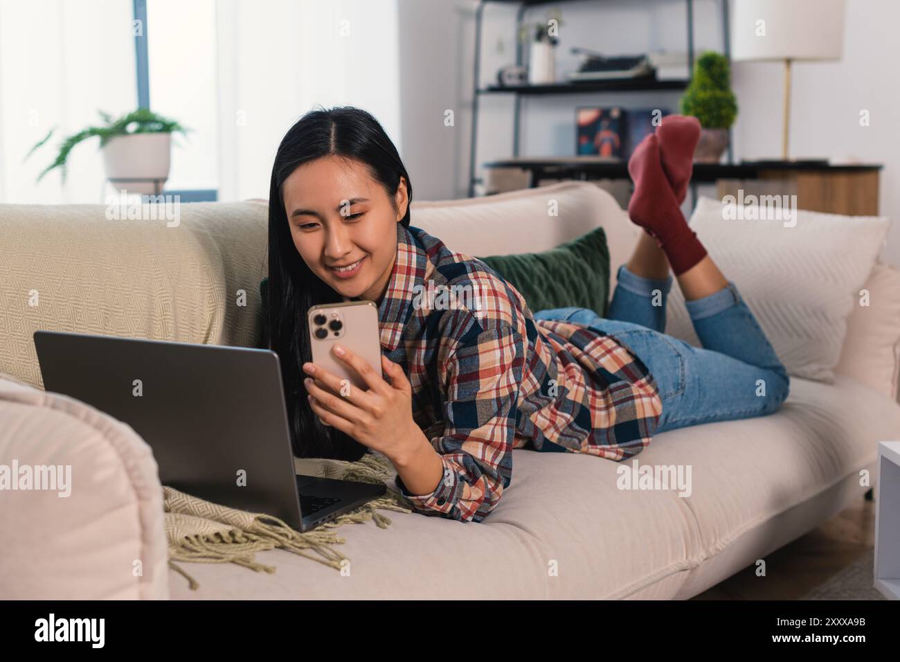 Giovane donna asiatica coreana generazione Z che lavora a casa e studia in remoto, imparando con un computer portatile e uno smartphone moderno il comodo salotto Foto Stock