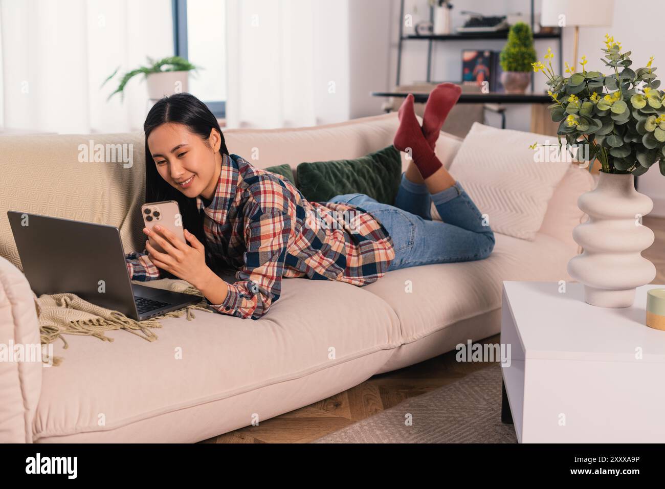 Giovane donna asiatica coreana generazione Z che lavora a casa e studia in remoto, imparando con un computer portatile e uno smartphone moderno il comodo salotto Foto Stock