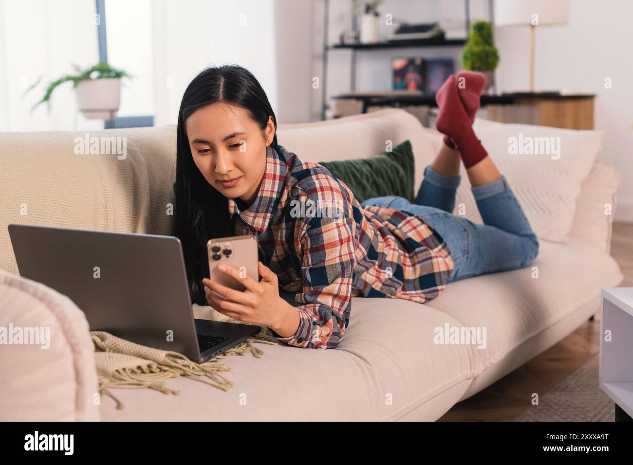 Giovane donna asiatica coreana generazione Z che lavora a casa e studia in remoto, imparando con un computer portatile e uno smartphone moderno il comodo salotto Foto Stock
