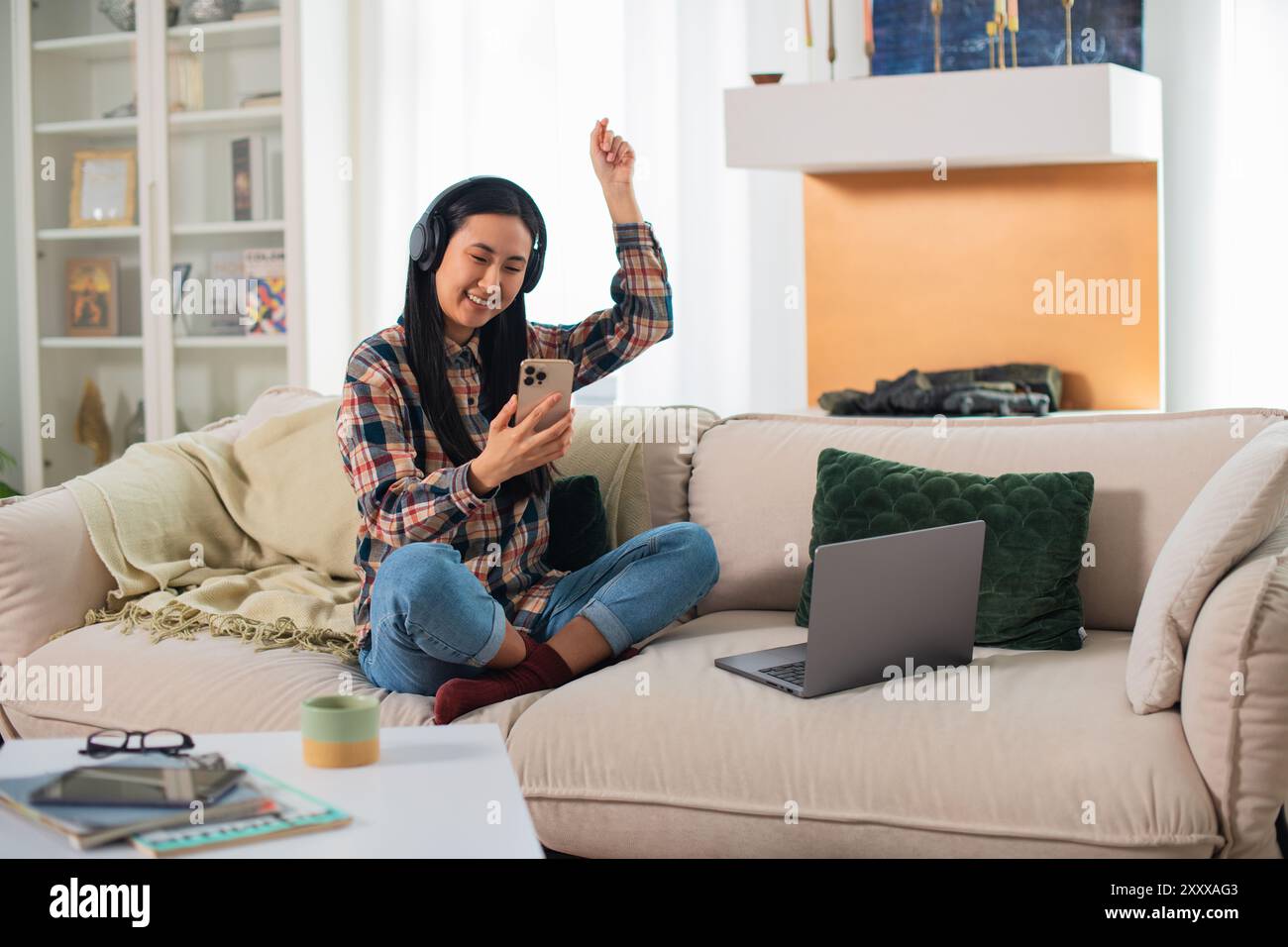 Giovane studentessa asiatica sorridente di generazione Z in abbigliamento casual con cuffie wireless che utilizzano su uno smartphone moderno nel confortevole soggiorno Foto Stock