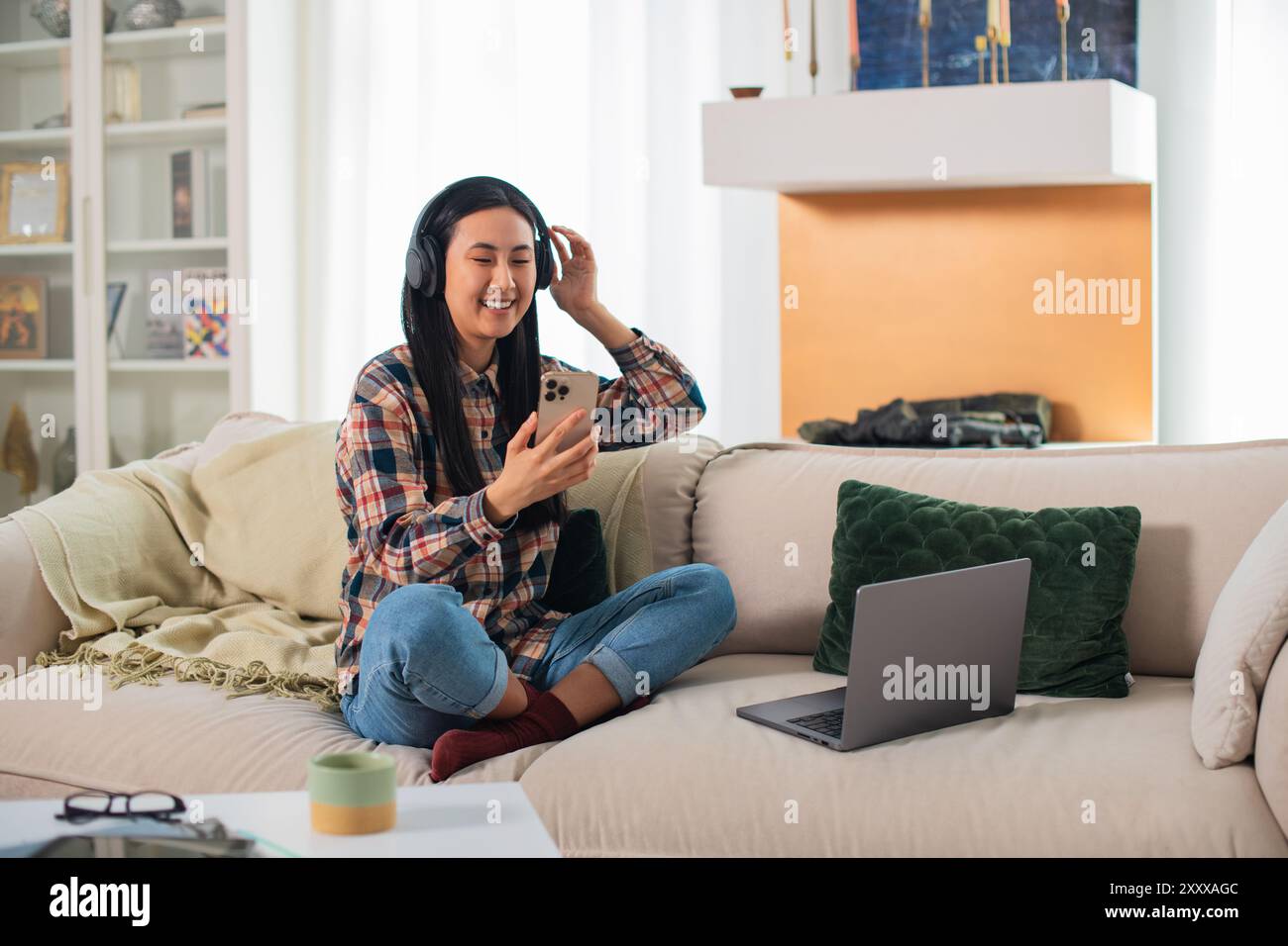 Giovane studentessa asiatica sorridente di generazione Z in abbigliamento casual con cuffie wireless che utilizzano su uno smartphone moderno nel confortevole soggiorno Foto Stock