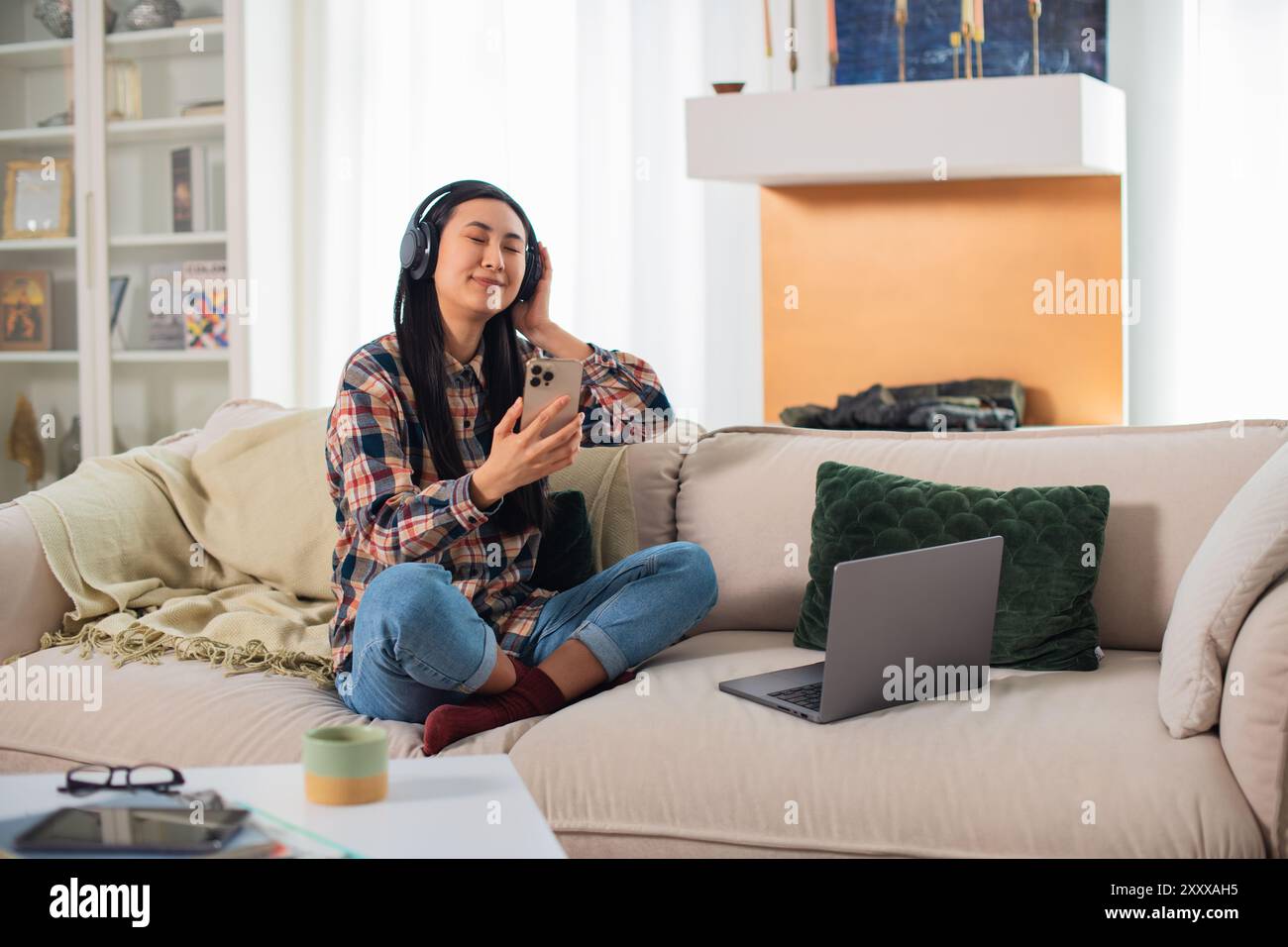 Giovane studentessa asiatica sorridente di generazione Z in abbigliamento casual con cuffie wireless che utilizzano su uno smartphone moderno nel confortevole soggiorno Foto Stock