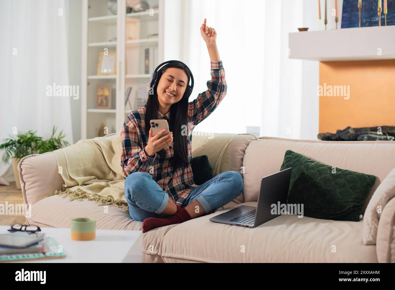 Giovane studentessa asiatica sorridente di generazione Z in abbigliamento casual con cuffie wireless che utilizzano su uno smartphone moderno nel confortevole soggiorno Foto Stock