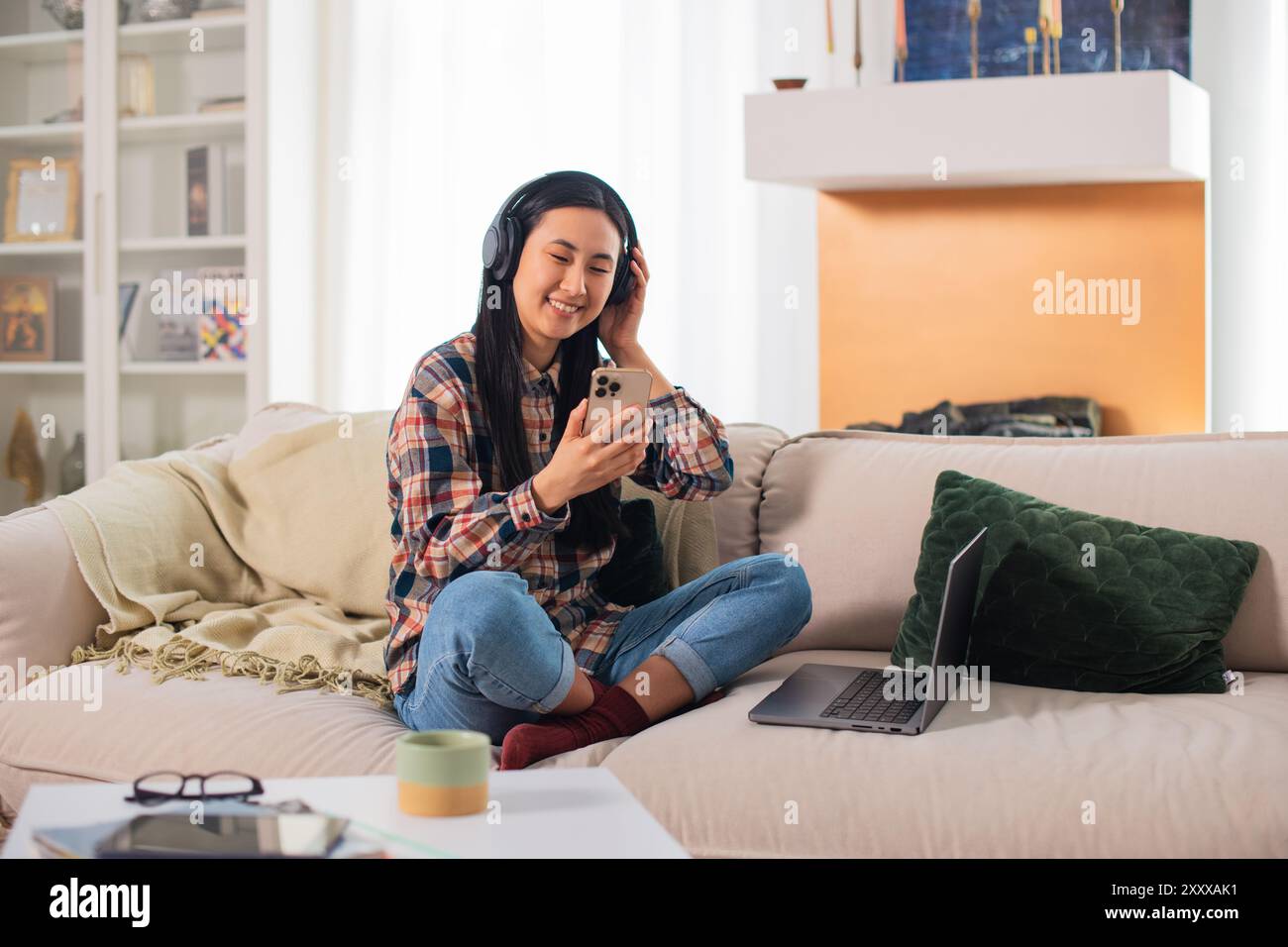 Giovane studentessa asiatica sorridente di generazione Z in abbigliamento casual con cuffie wireless che utilizzano su uno smartphone moderno nel confortevole soggiorno Foto Stock