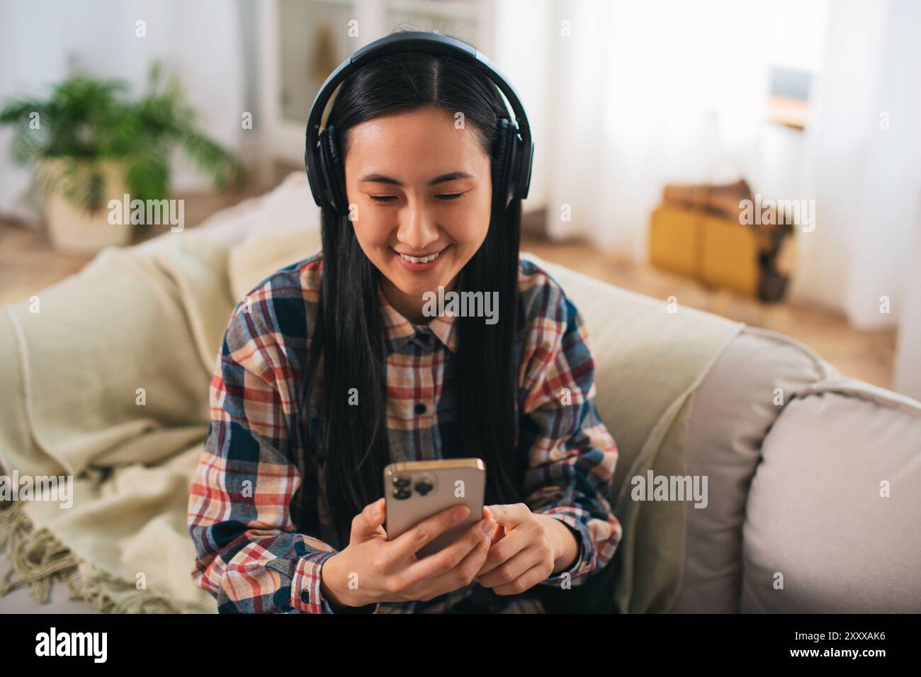 Giovane studentessa asiatica sorridente di generazione Z in abbigliamento casual con cuffie wireless che utilizzano su uno smartphone moderno nel confortevole soggiorno Foto Stock