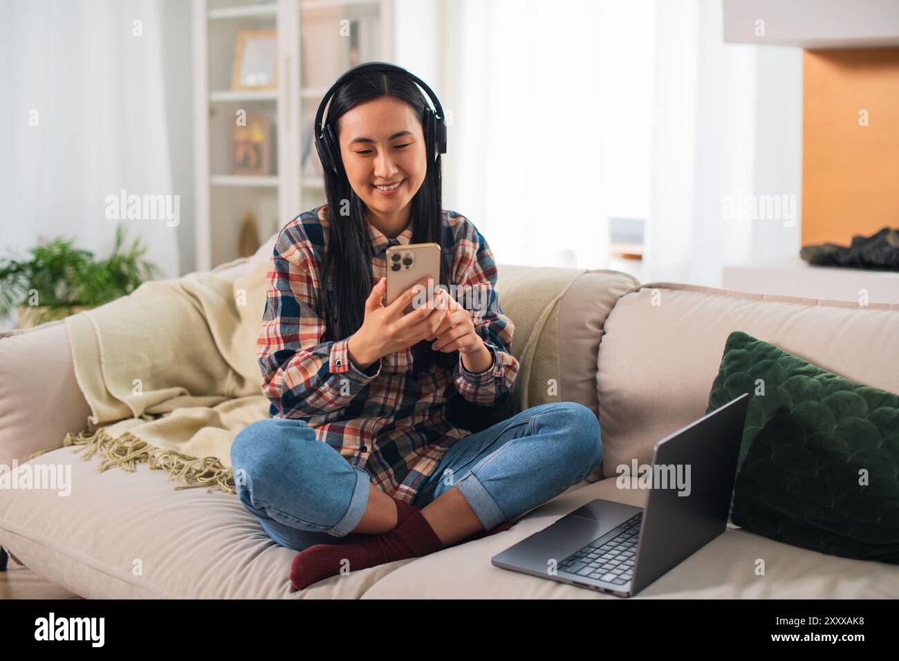 Giovane studentessa asiatica sorridente di generazione Z in abbigliamento casual con cuffie wireless che utilizzano su uno smartphone moderno nel confortevole soggiorno Foto Stock