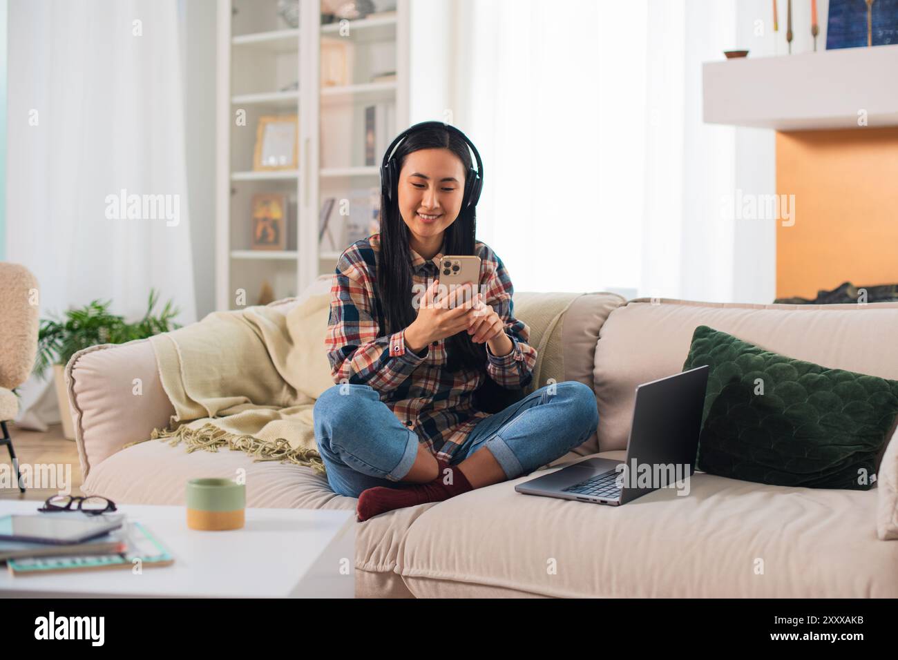 Giovane studentessa asiatica sorridente di generazione Z in abbigliamento casual con cuffie wireless che utilizzano su uno smartphone moderno nel confortevole soggiorno Foto Stock