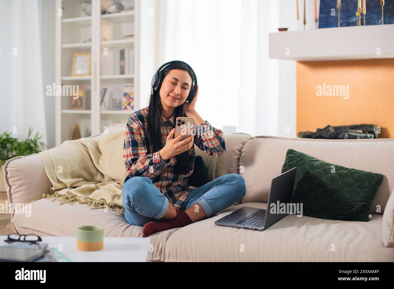 Giovane studentessa asiatica sorridente di generazione Z in abbigliamento casual con cuffie wireless che utilizzano su uno smartphone moderno nel confortevole soggiorno Foto Stock