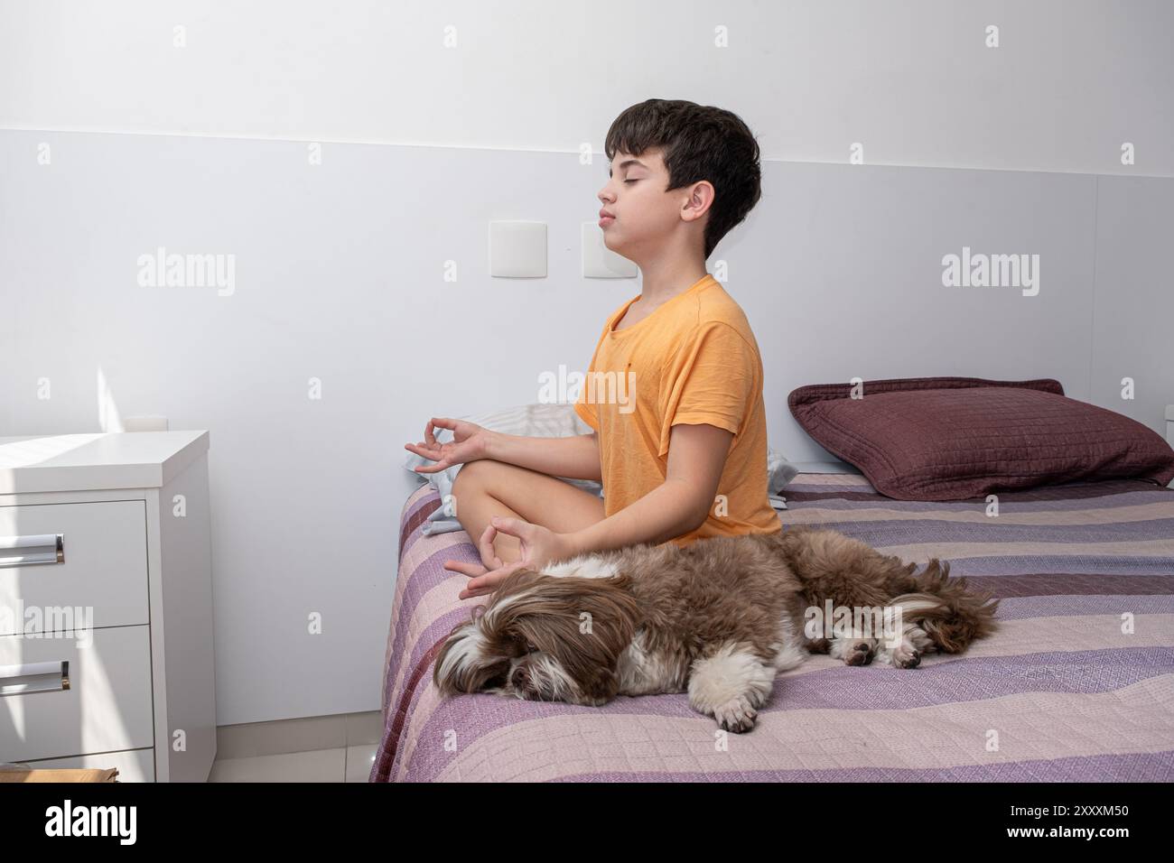 Ragazzo brasiliano di 11 anni seduto sul suo letto, praticando yoga accanto al suo cane 1. Foto Stock