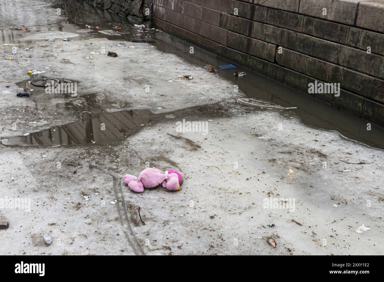 Spazzatura, una ruota da bici e un grande giocattolo rosa sul ghiaccio parzialmente fuso in un canale Foto Stock