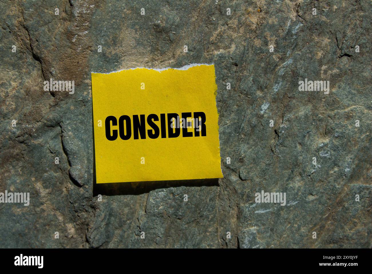 Considerare il messaggio scritto su un pezzo di carta strappato giallo con sfondo in pietra. Simbolo concettuale da considerare. Copia spazio. Foto Stock