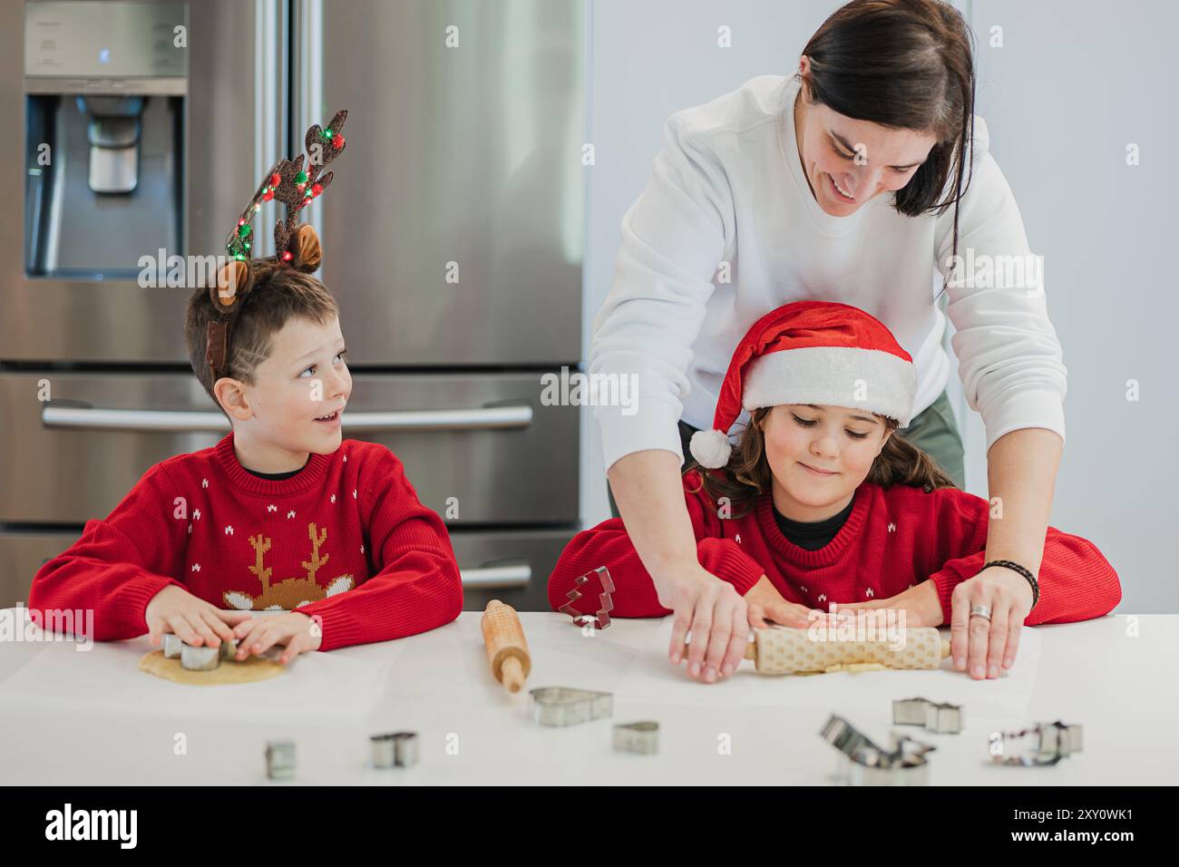 Una scena commovente di una madre che aiuta i suoi figli gemelli, vestiti con abiti festivi, stendere l'impasto per i biscotti di Natale in una moderna cucina Foto Stock