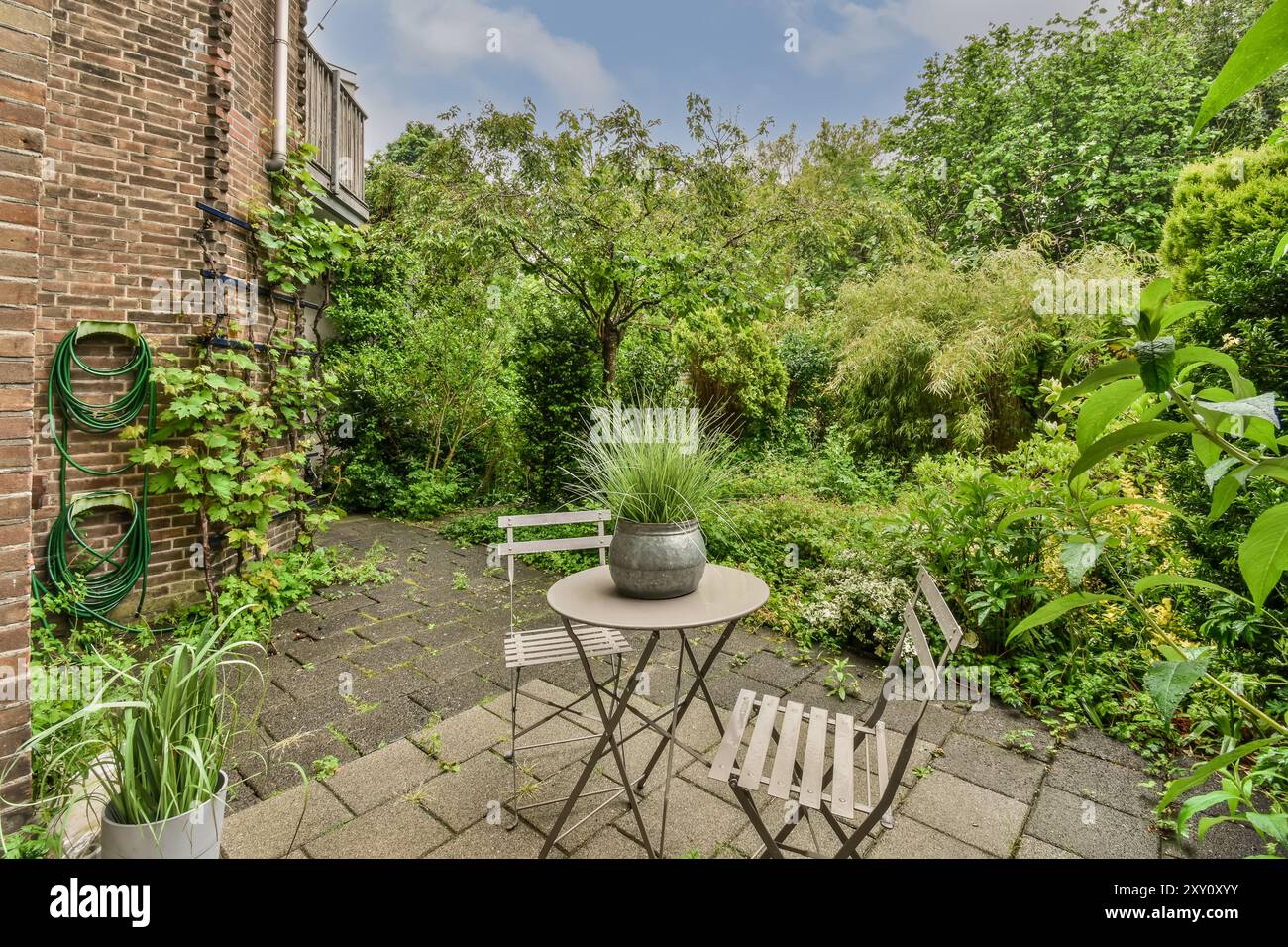 Un'accogliente area giardino esterna con tavolo da bistro e sedie adagiate su una parete di mattoni, circondate da lussureggiante vegetazione. Foto Stock