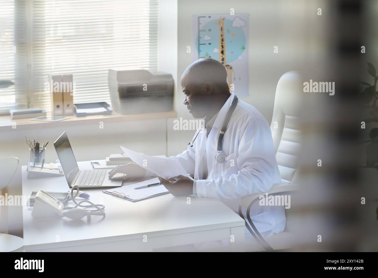 Medico professionista che analizza documenti in un ambiente d'ufficio con luce solare brillante che arriva attraverso le tende da finestra. I dintorni includono vari punti vendita Foto Stock