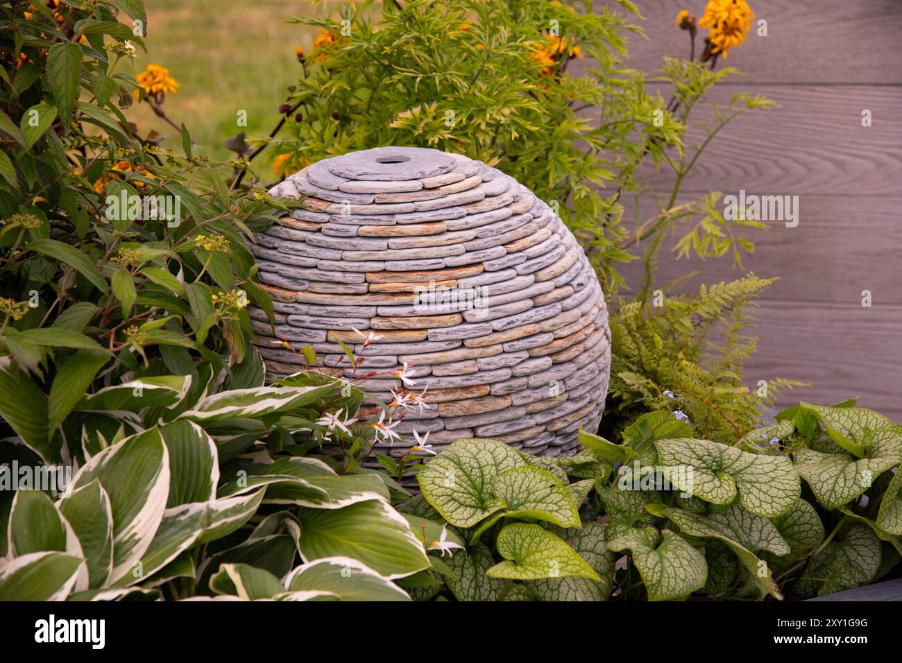 Una scultura a forma di sfera di pietra circondata da Hostas e Brunera. Foto Stock