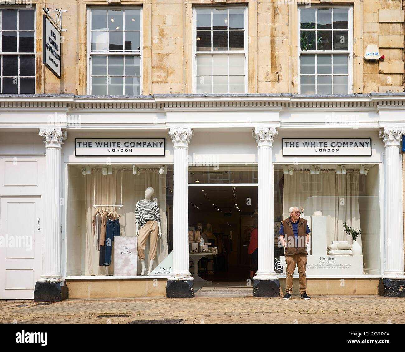 Un uomo sta aspettando, con le mani in tasca, fuori dal negozio di abbigliamento della White Company sulla strada pedonale di High Street, Stamford, Inghilterra. Foto Stock