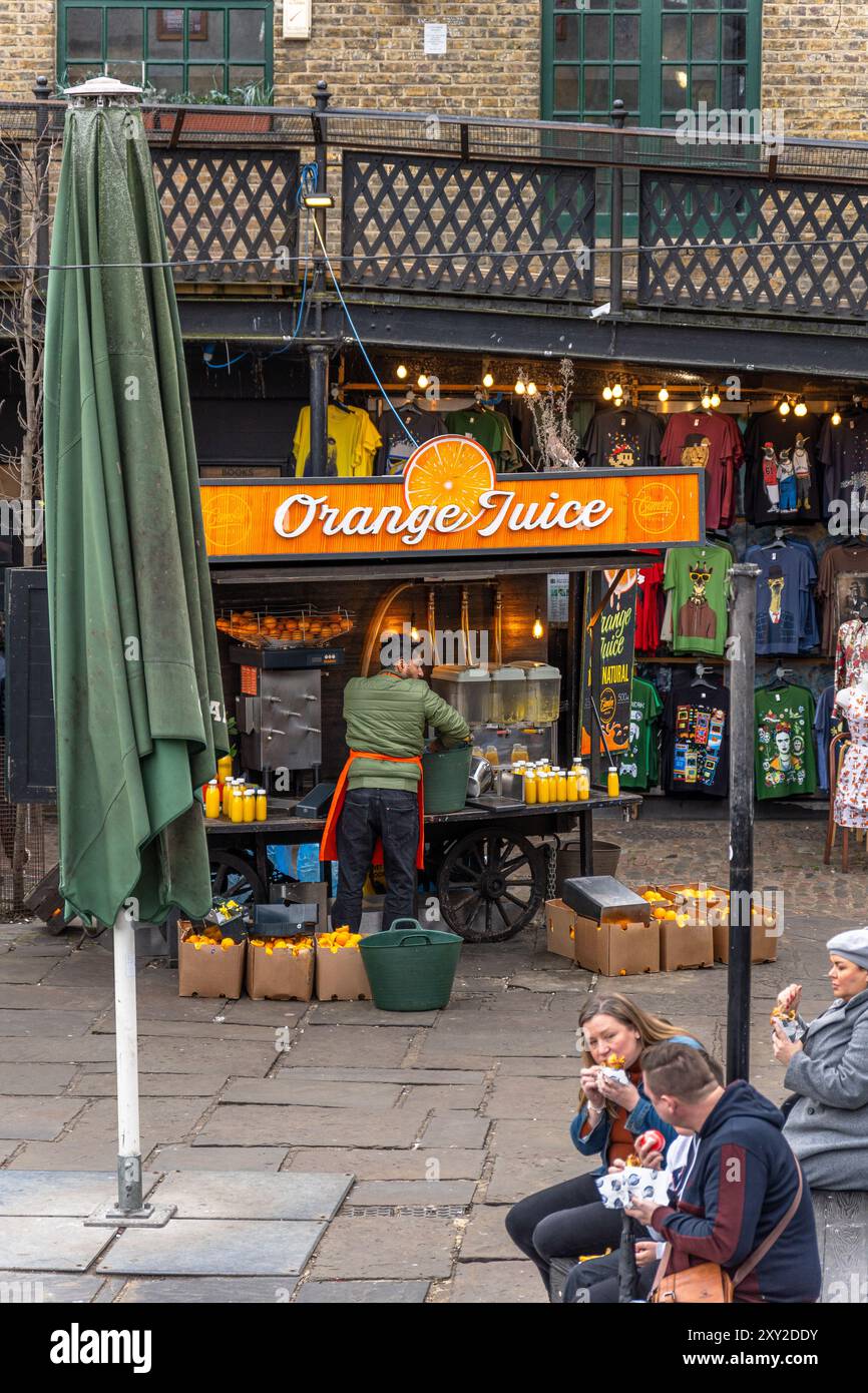 Il cameriere prepara le arance in una bancarella di succo d'arancia a Camden Market, West Yard, e i turisti che mangiano seduti accanto a un chiosco di t-shirt geek. Londra. Foto Stock