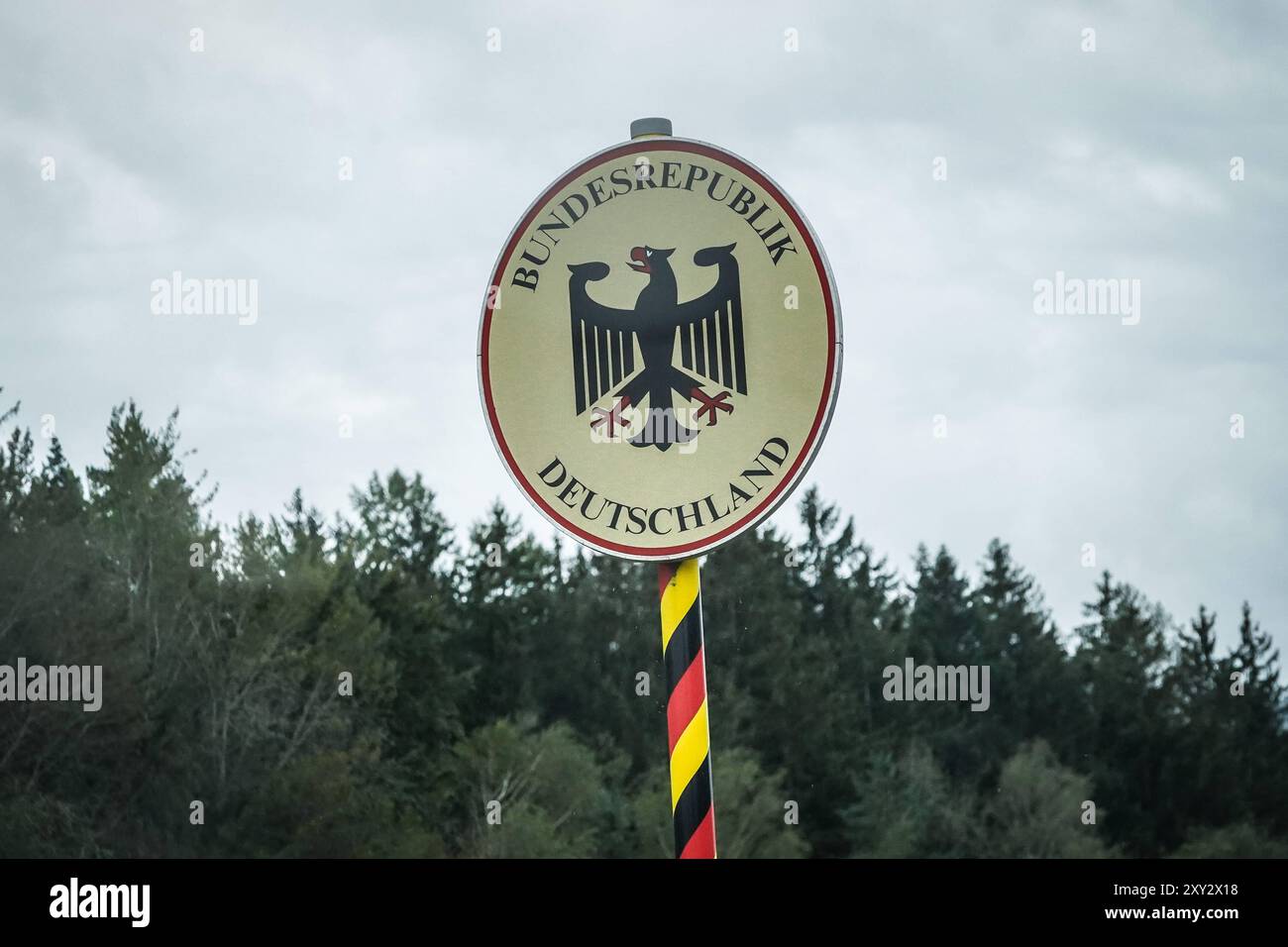 Grenzschild der Bundesrepublik Deutschland, Wappen mit Bundesadler, schwarz-rot-gelbe Markierung am Pfosten, bewaldete Landschaft im Hintergrund, wolkiger Himmel, Symbol für nationale Grenze, Grenzübertritt, Grenzkontrollen, nationale Sicherheit, Grenzpolitik, Einreisebestimmungen, deutsch-polnische Grenze, EU-Außengrenze, Migrationsfragen, nationale Souveränität, internationale Beziehungen, Grenzschutz. *** Segno di confine della Repubblica Federale di Germania, stemma con aquila federale, contrassegno nero rosso giallo sul palo, paesaggio boscoso sullo sfondo, cielo nuvoloso, simbolo per la nazionale b Foto Stock