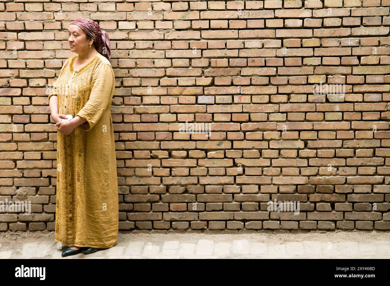 Una donna uigura in piedi accanto a un muro marrone in mattoni a Kashgar, Xinjiang, Cina. Foto Stock