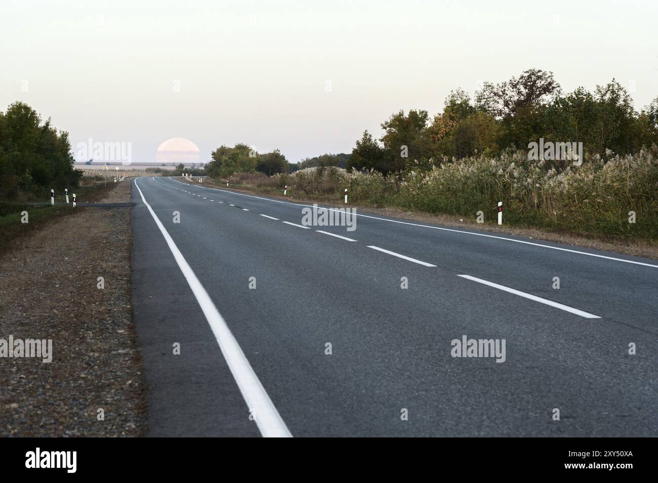 Un'enorme luna piena al mattino presto che lascia l'orizzonte sopra la strada asfaltata. Foto vera Foto Stock
