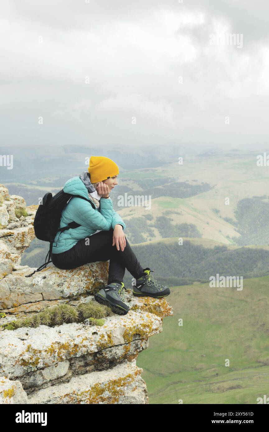 Giovane donna con uno zaino che si siede sul bordo di una roccia e guarda il cielo con le nuvole Foto Stock