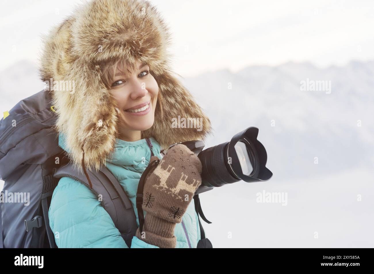 Ritratto ravvicinato di una ragazza sorridente viaggiatrice felice con un cappello di pelliccia grande e guanti in maglia con una macchina fotografica in mano. Zaino sul retro. Montagne i Foto Stock