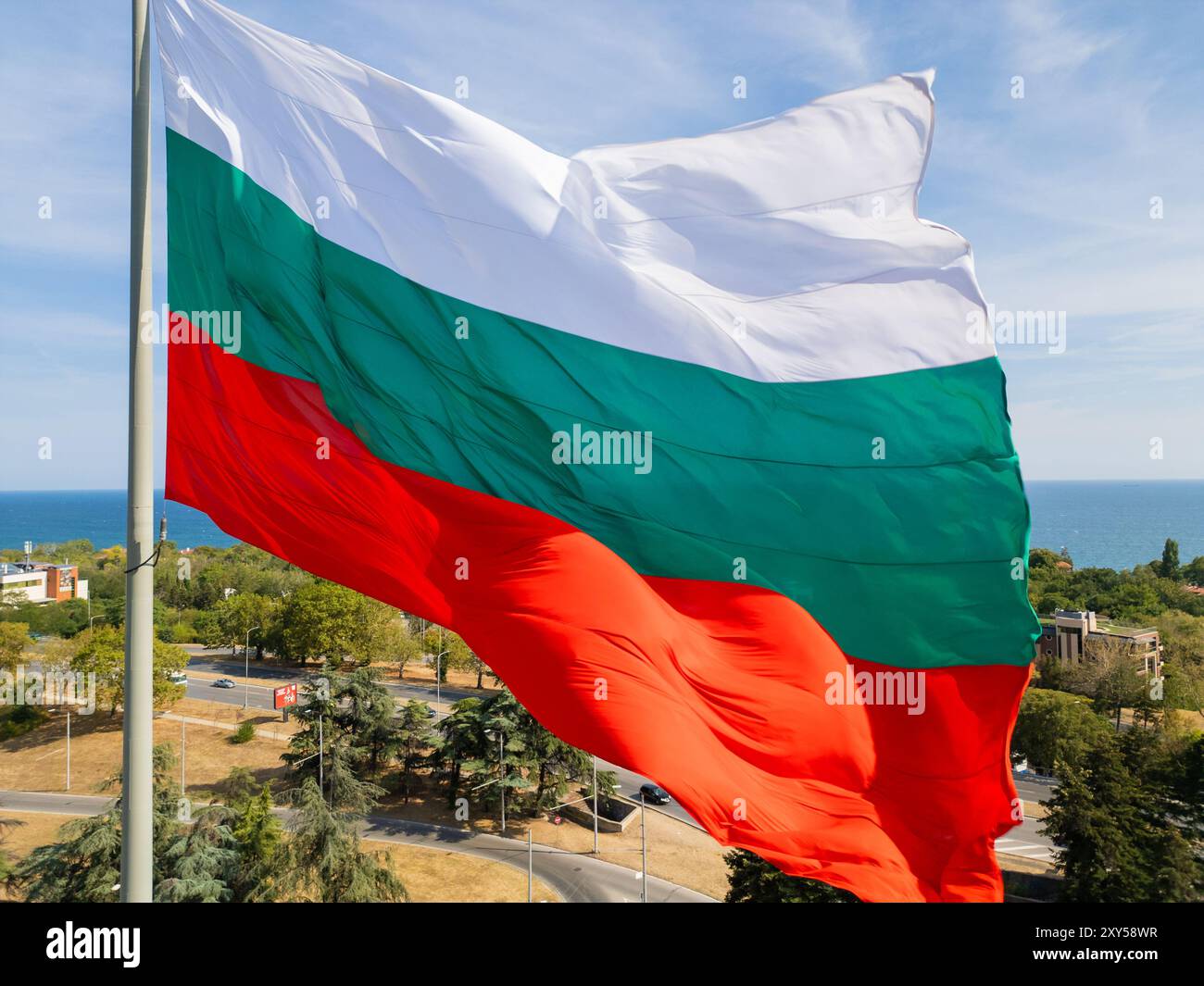 La bandiera bulgara ondeggia orgogliosamente sopra una città costiera, con il vasto mare e il verde paesaggio sullo sfondo. Un simbolo di orgoglio nazionale e.. Foto Stock