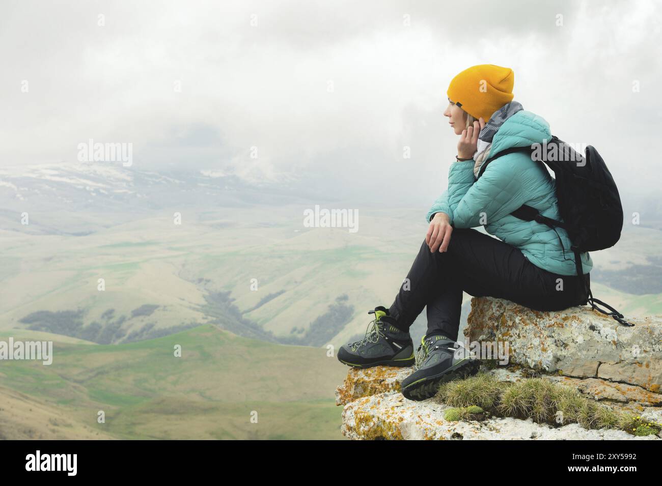 Giovane donna con uno zaino che si siede sul bordo di una roccia e guarda il cielo con le nuvole Foto Stock