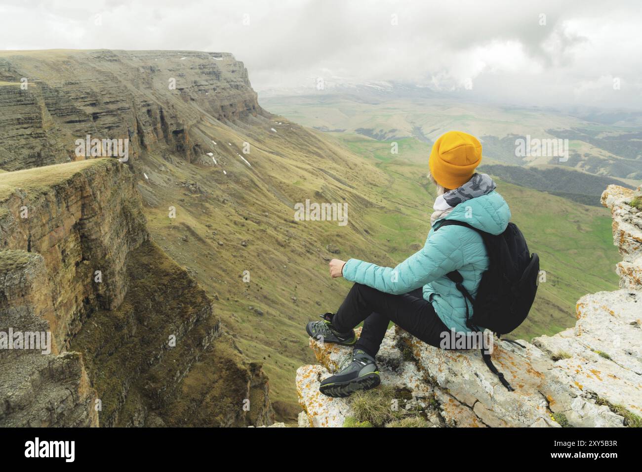 Giovane donna con uno zaino che si siede sul bordo di una roccia e guarda il cielo con le nuvole Foto Stock