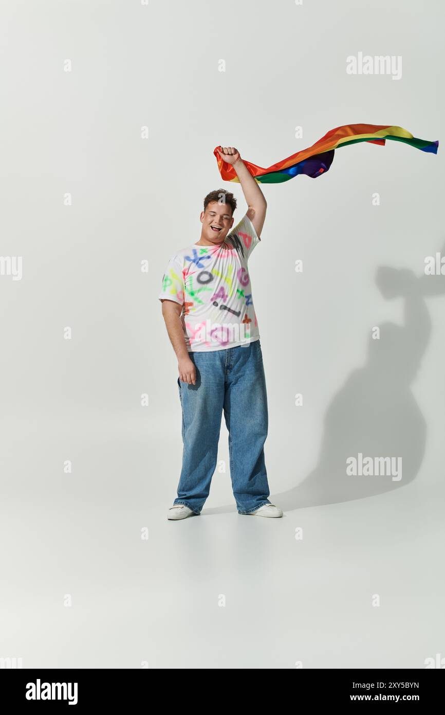 Una persona queer con una t-shirt colorata regge una bandiera arcobaleno e sorride. Foto Stock