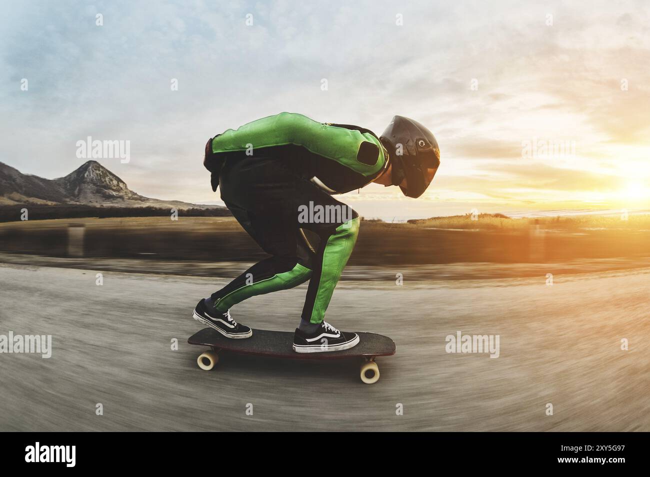 Un giovane con casco integrale e una tuta in pelle in uno speciale rack, cavalca un longboard sul tramonto afsaltu sulle montagne sullo sfondo e sul bellissimo sk Foto Stock Un giovane con casco integrale e una tuta in pelle in uno speciale rack, cavalca un longboard sul tramonto afsaltu sulle montagne sullo sfondo e sul bellissimo sk Foto Stock