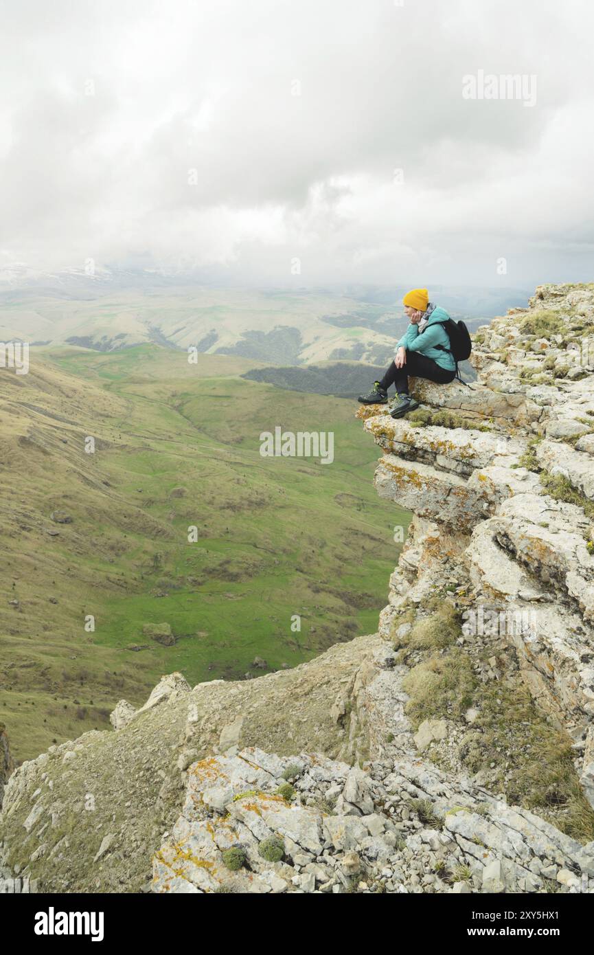 Giovane donna con uno zaino che si siede sul bordo di una roccia e guarda il cielo con le nuvole Foto Stock
