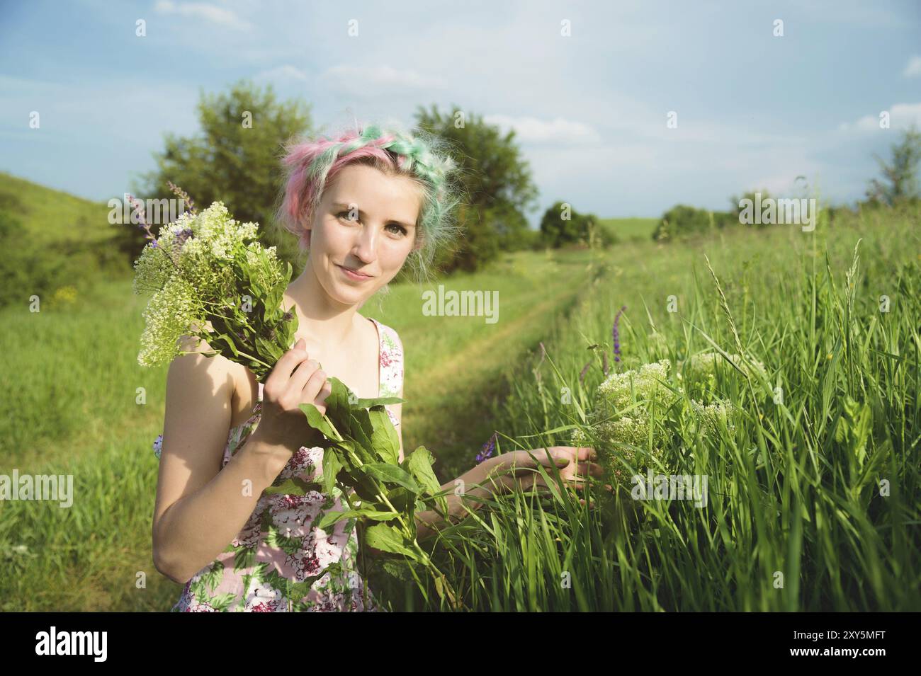 Ritratto di una giovane ragazza felice e carina con capelli multicolori raccoglie fiori accanto a una strada di campagna al tramonto. Il concetto di armonia umana con la n Foto Stock