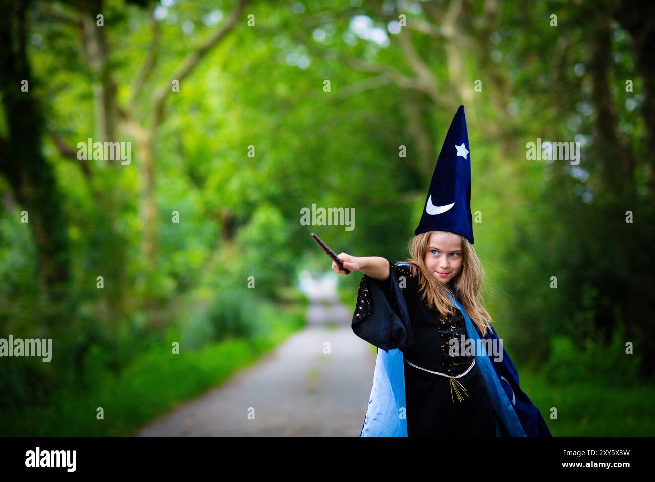 Bambina bionda caucasica in costume blu mago con mantello e cappello, che lancia un incantesimo con bacchetta magica sullo sfondo della foresta all'aperto. Foto Stock