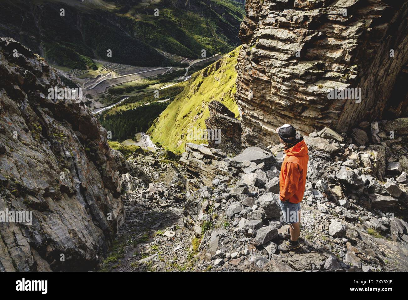 Il ritratto di un escursionista maschile in pantaloncini corti e un berretto dal retro è in piedi su alte rocce ai margini di una scogliera sullo sfondo di una valle verde W Foto Stock