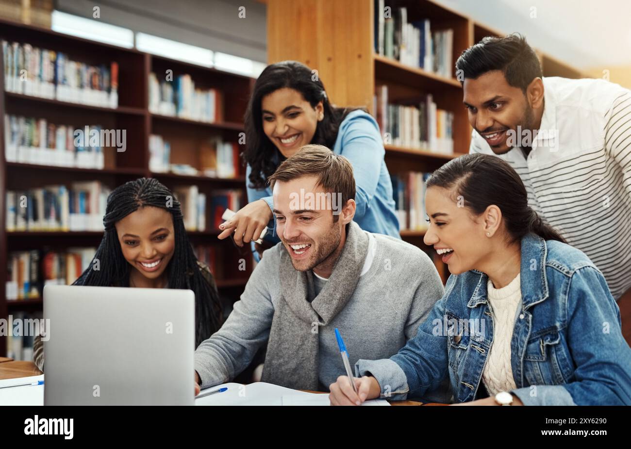Università, studenti e ridendo di laptop in biblioteca per progetti di ricerca, meme divertenti e compiti creativi. Gruppo di studio sulla diversità, libri e. Foto Stock