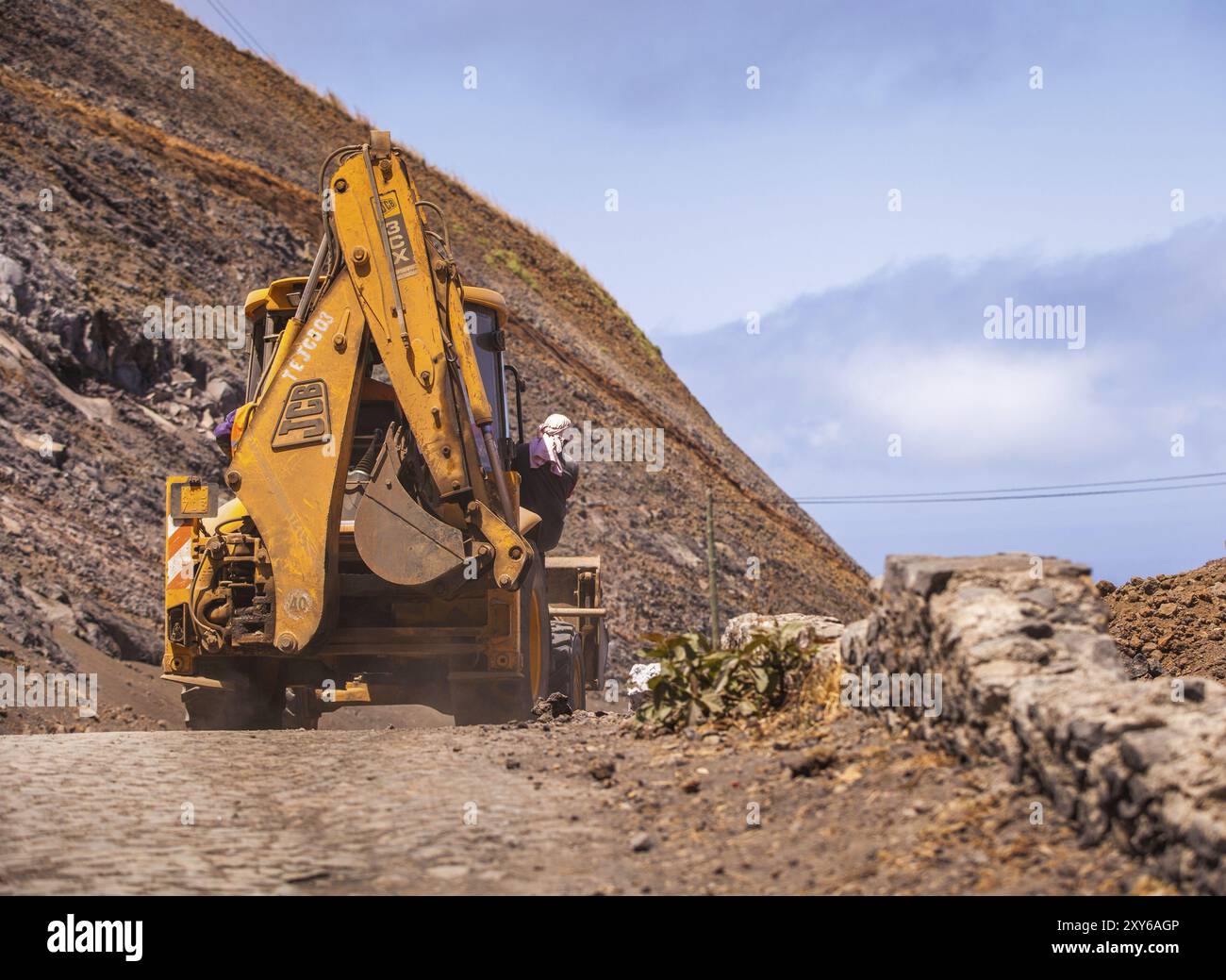 Escavatore giallo che viaggia lungo una strada polverosa e un uomo aggrappato al suo fianco Foto Stock