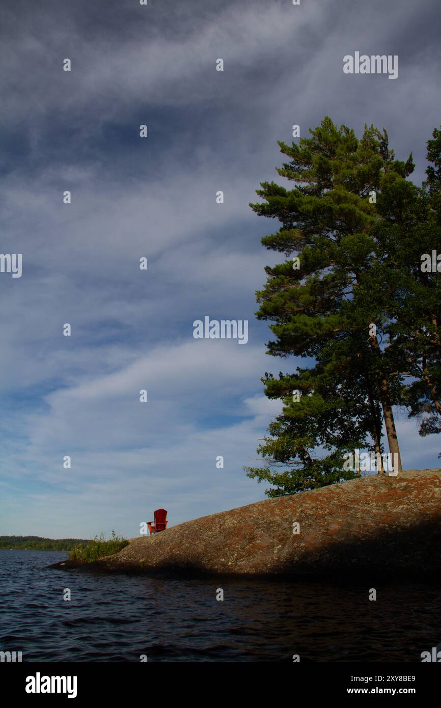 Una sedia rossa vuota Adirondack su una costa rocciosa con alcuni alberi lungo il lago, Muskoka Lake, Ontario, Canada Foto Stock